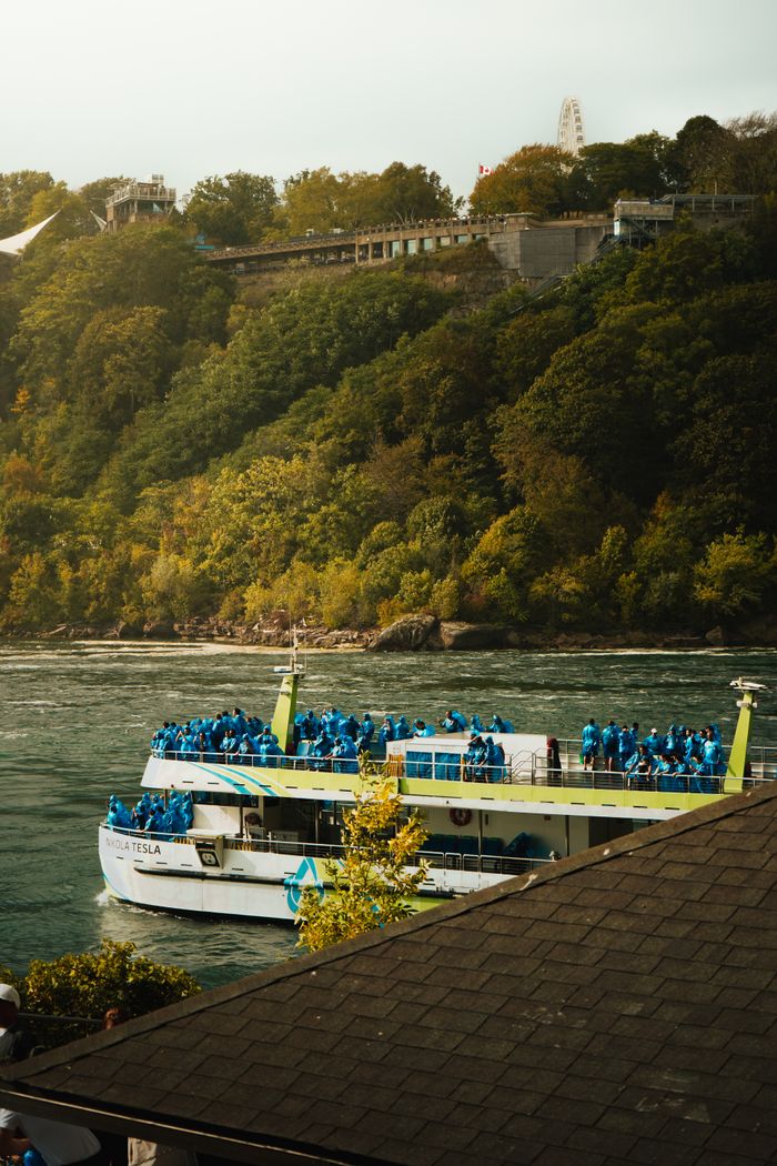 Tourist boat at Niagara River with lush green hillside and distant ferris wheel.